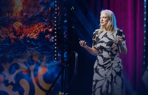 "Speaker presenting on stage in a floral dress at a legal ops conference, with colorful stage lighting in the background"